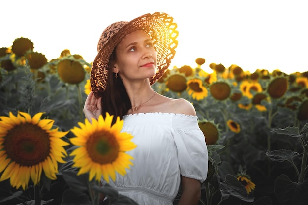 Una giovane ragazza con un vestito bianco e un cappello in un campo di girasoli al tramonto Ritratto di una donna dalla figura snella su uno sfondo di fiori gialli