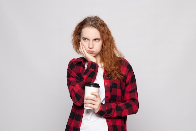 Una giovane ragazza con una camicia a quadri tiene una tazza di caffè su uno sfondo chiaro