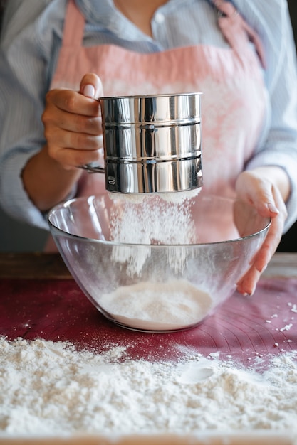 Una giovane ragazza cuoca con un grembiule setaccia la farina di frumento bianco in una ciotola di vetro. Preparazione dell'impasto per la cottura del khachapuri. Setaccio di ferro per farina.