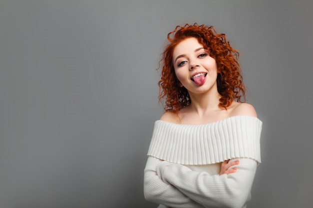 Una giovane ragazza dai capelli rossi dai capelli ricci mostra scherzosamente la lingua nella fotocamera con le mani giunte sul petto.