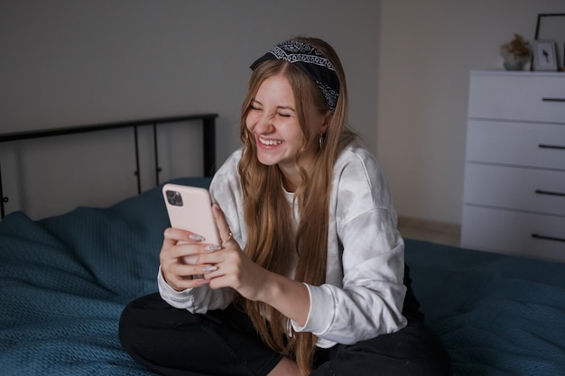 Una giovane ragazza di 20 anni si siede su un letto nel suo soggiorno tiene un telefono in mano e ride
