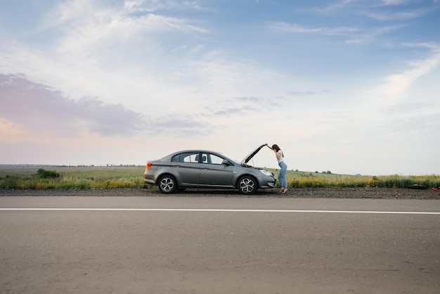 Una giovane ragazza è in piedi vicino a un'auto in panne nel mezzo dell'autostrada durante il tramonto e cerca di chiamare aiuto al telefono. Riparazione e riparazione dell'auto. In attesa di aiuto.