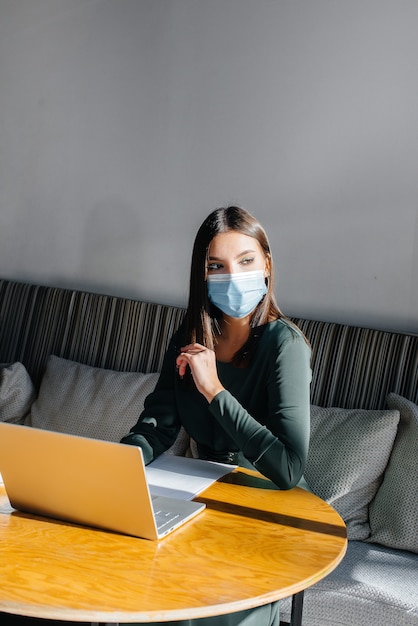 Una giovane ragazza è seduta in un bar con indosso una maschera e lavora al computer.