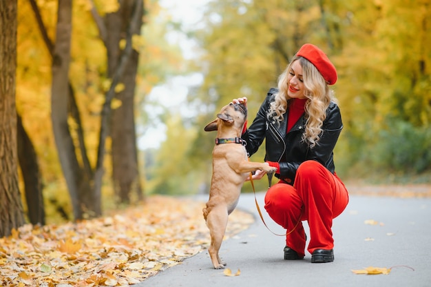 Una giovane ragazza elegante con lunghi capelli chiari in occhiali soleggiati va a fare una passeggiata con un piccolo cagnolino medio un carlino del bulldog francese in un parco in primavera in autunno