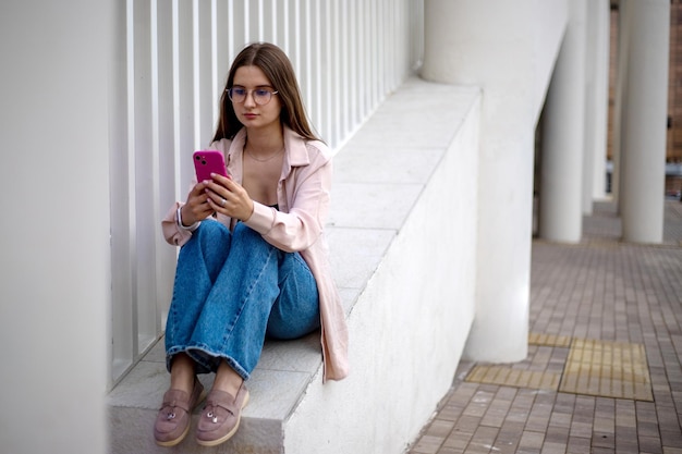 una giovane ragazza in jeans, una maglietta con un telefono si siede per strada e sorride