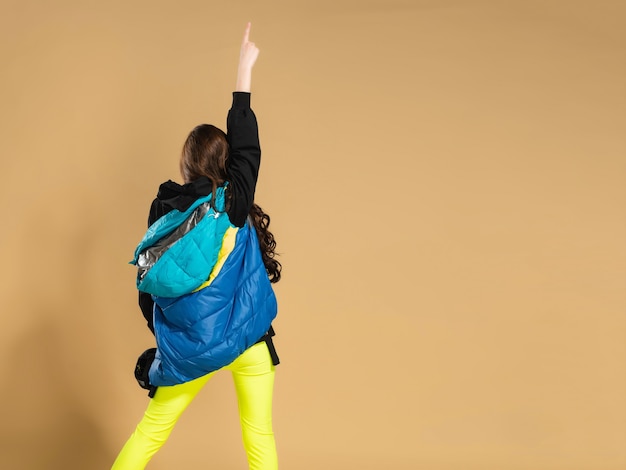 Una giovane ragazza in leggings gialli e un caldo gilet alza la mano con l'indice rivolto verso l'alto e volta la schiena contro un arancio pastello.