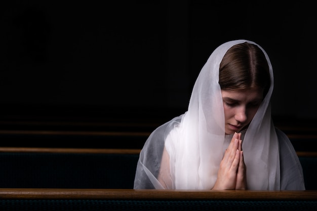 Una giovane ragazza modesta con un fazzoletto in testa è seduta in chiesa e prega. La religione, la preghiera, il culto