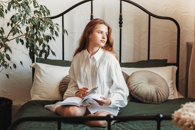 Una giovane ragazza romantica con i capelli lunghi si siede su un letto con un libro. Leggere a letto