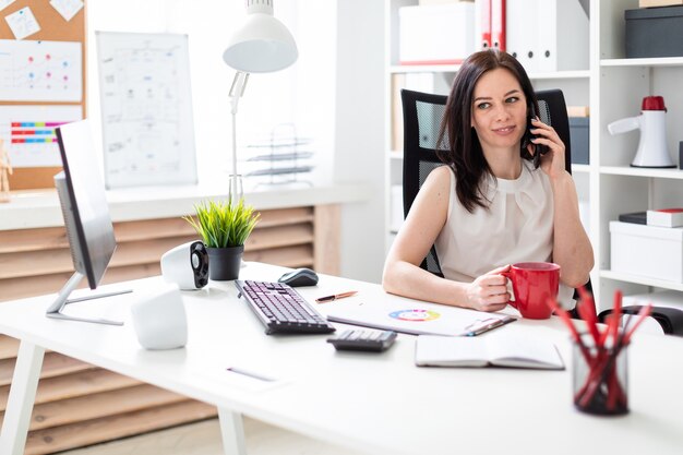 Una giovane ragazza seduta in ufficio alla scrivania del computer, parlando al telefono e in possesso di una tazza rossa.