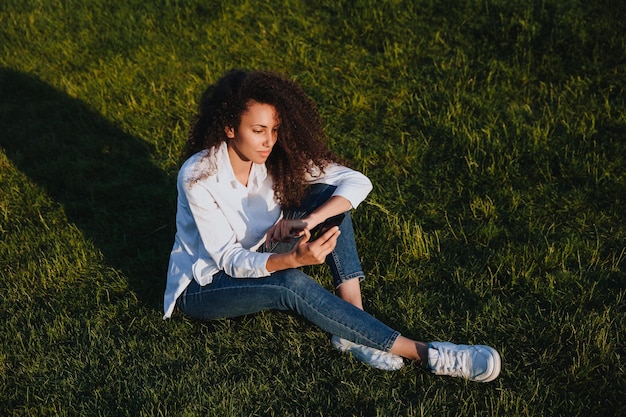 Una giovane ragazza si siede sull'erba verde nel parco e utilizza i social network utilizzando un telefono cellulare