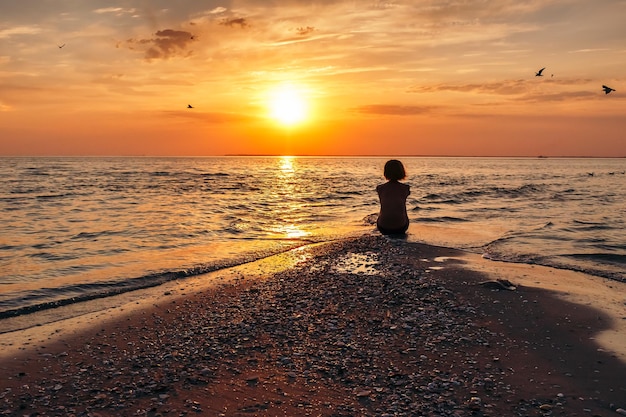 Una giovane ragazza si siede sulla sabbia e guarda le piccole onde del mare e il tramonto arancione Silhouette di una ragazza sulla spiaggia Atmosfera calma di una sera d'estate Vista posteriore