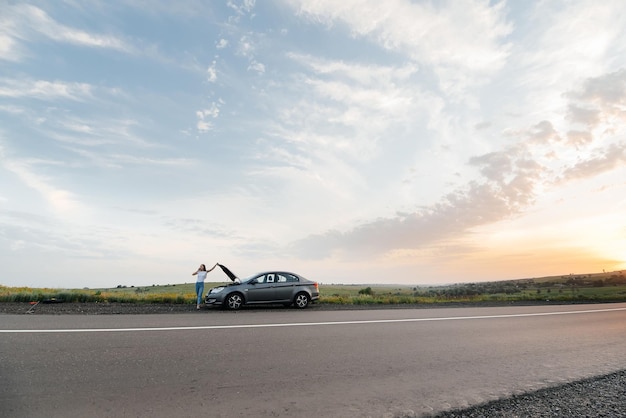 Una giovane ragazza si trova vicino a un'auto in panne in mezzo all'autostrada durante il tramonto e cerca di chiamare aiuto al telefono In attesa di aiuto Servizio auto Guasto auto su strada