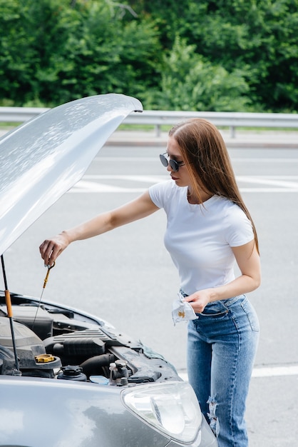 Una giovane ragazza si trova vicino a un'auto in panne in mezzo all'autostrada e controlla il livello dell'olio nel motore. Guasto e guasto dell'auto. In attesa di aiuto.