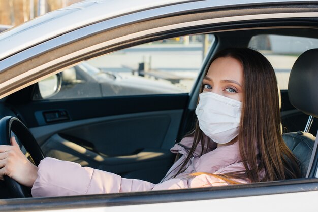 Una giovane ragazza siede al volante in macchina nella maschera durante la pandemia globale e il coronavirus. Quarantena.