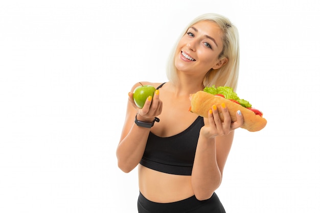 Una giovane ragazza sportiva con i capelli biondi in un top sportivo nero e leggings neri tiene una mela verde e un panino