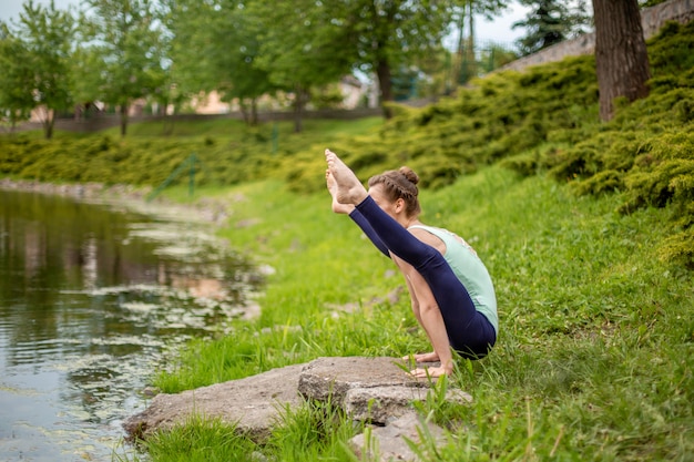 Una giovane ragazza sportiva pratica yoga su un prato verde vicino al fiume, yoga assans postura. Meditazione e unità con la natura