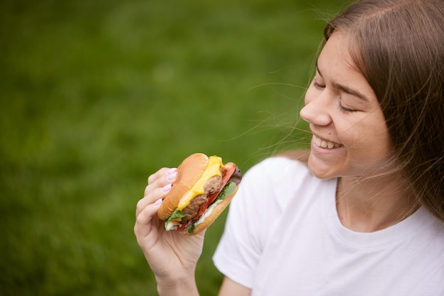 Una giovane ragazza tira fuori un hamburger da un sacchetto di carta seduto sull'erba verde il concetto di cibo di strada per la consegna del cibo