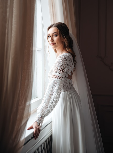 Una giovane sposa in un bellissimo abito di pizzo boho sta alla finestra.