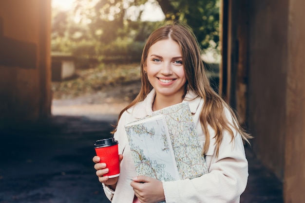 Una giovane studentessa cammina per la città con una mappa e tiene una tazza di caffè.