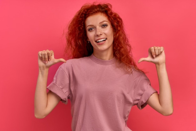 Una giovane studentessa dai capelli rossi allegra punta le dita su se stessa, il che significa una buona scelta isolata su sfondo rosa. Il concetto di raccomandazioni e scelte. Spazio pubblicitario