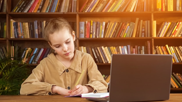 Una giovane studentessa in giacca marrone scrive lezioni su un quaderno di carta con una matita nella mano sinistra al computer portatile contro una grande libreria di legno in biblioteca