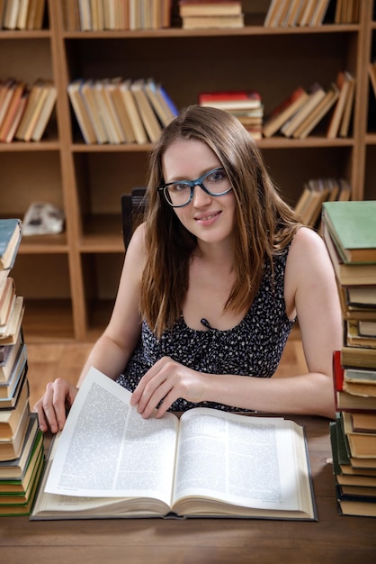 Una giovane studentessa legge un libro di testo seduto a un tavolo con molti libri in biblioteca