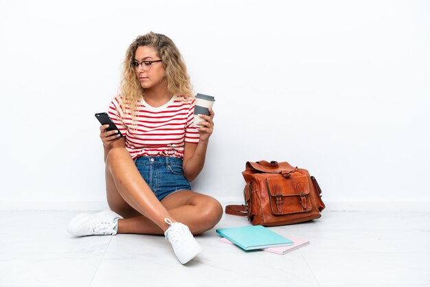 Una giovane studentessa seduta sul pavimento che tiene il caffè da portare via e un cellulare