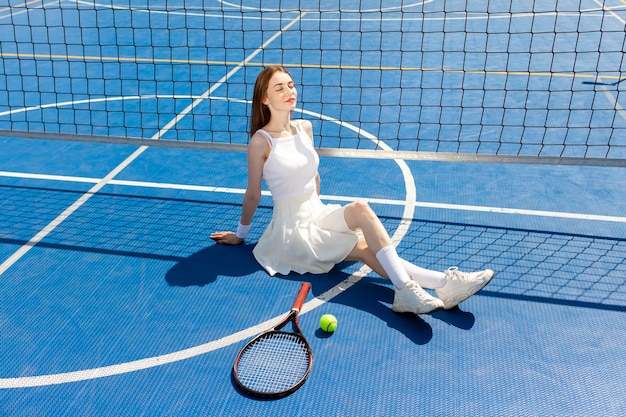 una giovane tennista in uniforme bianca con una racchetta si siede e si riposa sul campo blu all'aperto