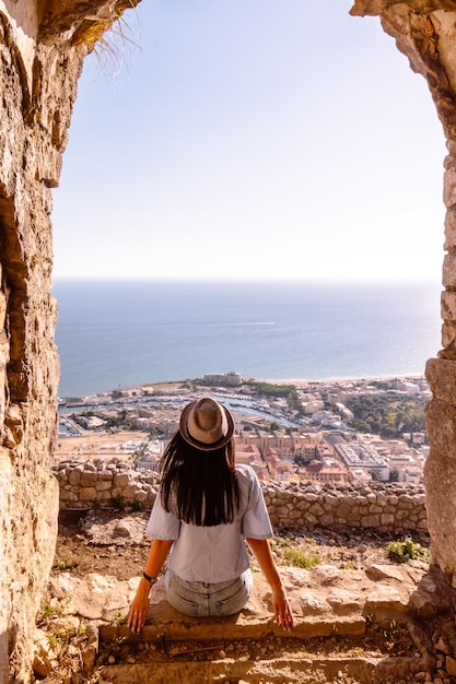 una giovane turista con un cappello si siede in cima a una montagna e guarda il mare e il porto