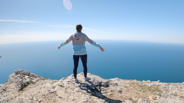 Una giovane viaggiatrice allarga le braccia ai lati e guarda il mare sullo scoglio. Ricreazione attiva e concetto di viaggio. La telecamera si muove intorno a una donna, 4K UHD.