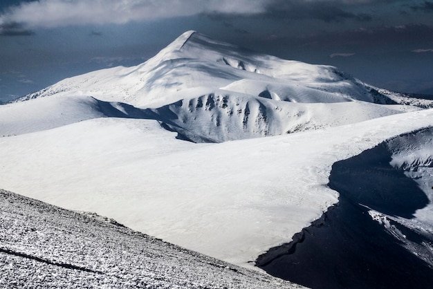 Una grande montagna rocciosa bianca con un couloir scuro è ricoperta di neve tra le nuvole nebbiose prima della bufera di neve invernale Hoverla