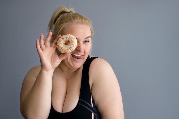 Una grassa donna bionda obesa in abiti sportivi chiude un occhio con una ciambella