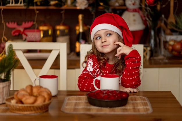 Una graziosa bambina con un maglione rosso e un cappello di Babbo Natale è in cucina a bere il tè con una torta e ad aspettare il nuovo anno o il Natale
