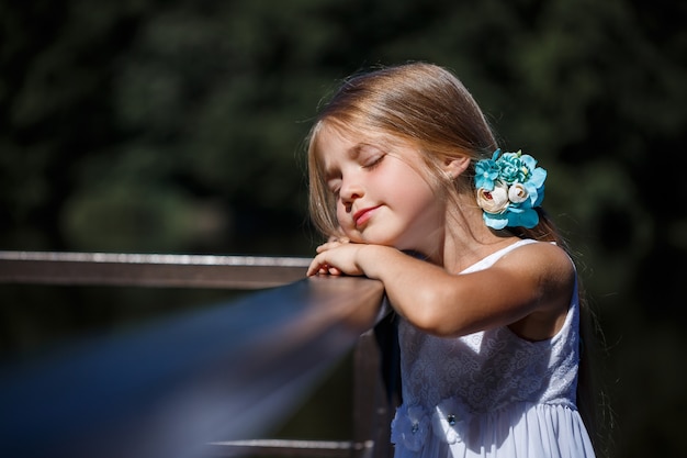 Una graziosa bambina di 6 o 7 anni con lunghi capelli biondi, è vestita con un abito estivo da balletto, è in piedi sullo sfondo del fiume