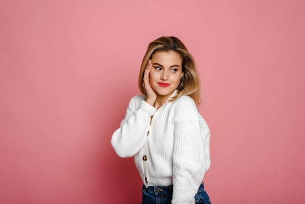 Una graziosa modella con un maglione bianco sorride e guarda la telecamera su uno sfondo rosa. Tenerezza e giovinezza.