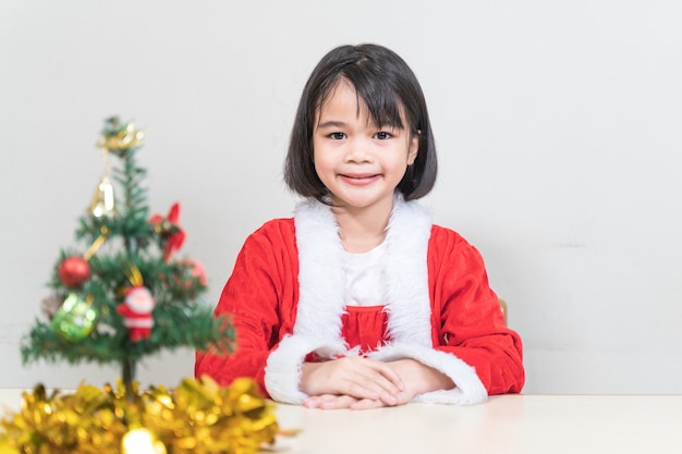 Una graziosa ragazzina asiatica con un vestito rosso da Babbo Natale che decora l'albero di Natale alla vigilia di Natale