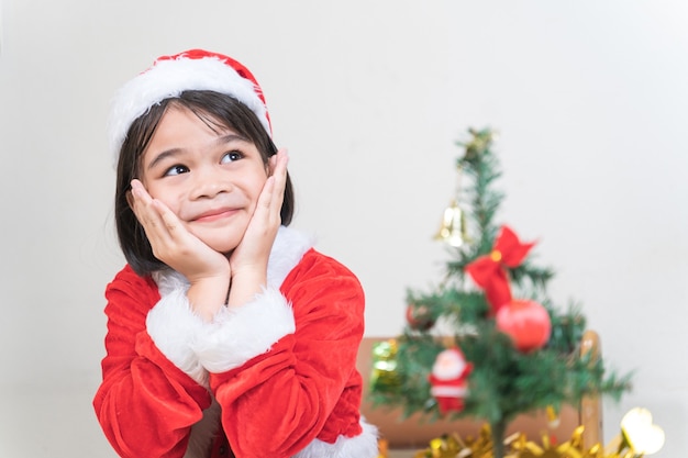 Una graziosa ragazzina asiatica con un vestito rosso da Babbo Natale che decora l'albero di Natale alla vigilia di Natale