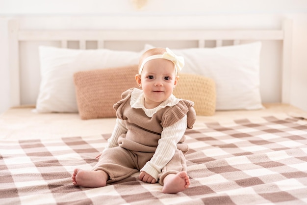 Una graziosa ragazzina europea con gli occhi marroni in un abito lavorato a maglia beige è seduta sul letto a casa sorridendo un ritratto di primo piano di un bambino