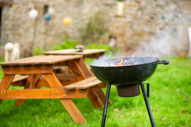 Una griglia rotonda fumante sullo sfondo di tavoli in legno con panche Preparazione per un picnic all'aperto