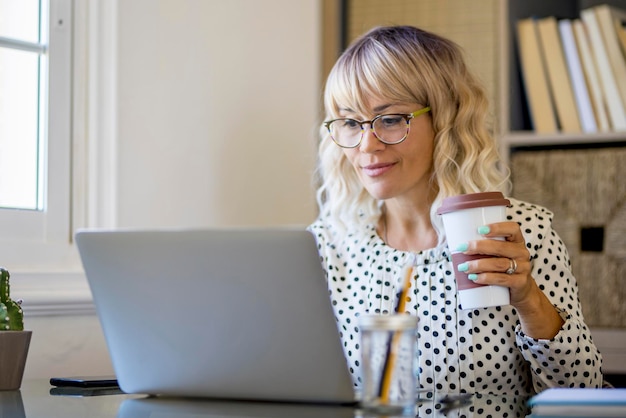 Una lavoratrice felice di successo che scrive sul laptop e beve caffè alla scrivania Home office freelance stile di vita occupato Donna d'affari online che utilizza il computer Attraente imprenditore di mezza età