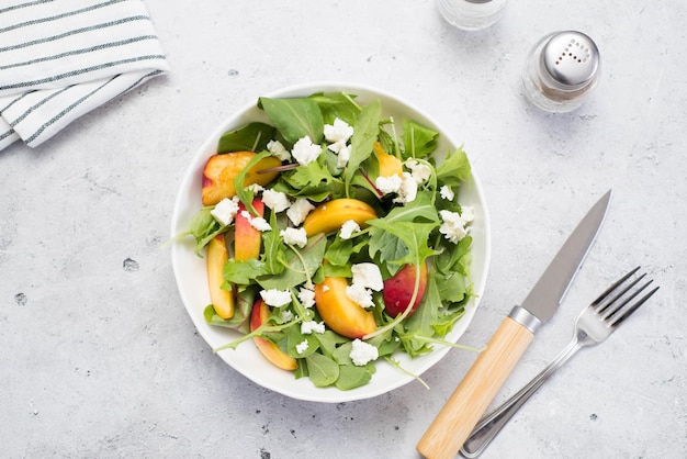 Una leggera insalata estiva di pesche rucolla e formaggio cucchiaio e coltello su uno sfondo grigio chiaro