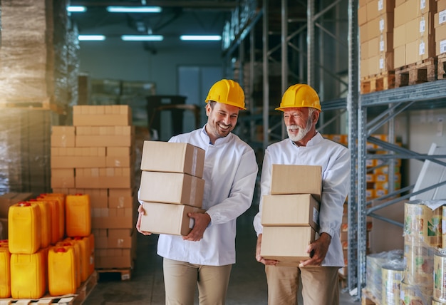 Una lunghezza di tre quarti di due impiegati sorridenti di stoccaggio caucasici in uniformi bianche e con caschi gialli che trasportano scatole in deposito.