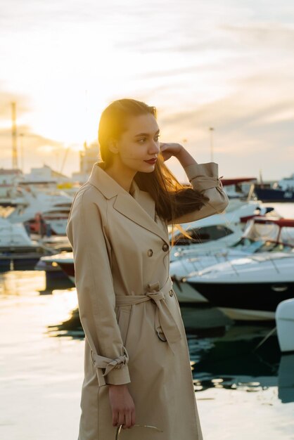 Una lussuosa giovane donna con i capelli lunghi in un cappotto beige alla moda è in piedi nel porto di sera e sta aspettando la sua nave