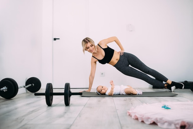 Una madre bionda sta facendo fitness con il suo bambino sul pavimento nella stanza Concetto di salute e recupero dopo il parto