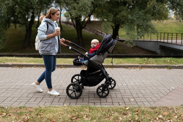 Una madre di un anno cammina con il suo bambino in un passeggino nel parco mentre beve il caffè da una tazza