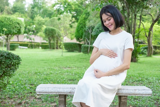 Una madre incinta in abiti casual bianchi si siede a guardare il suo bambino sul prato del parco.