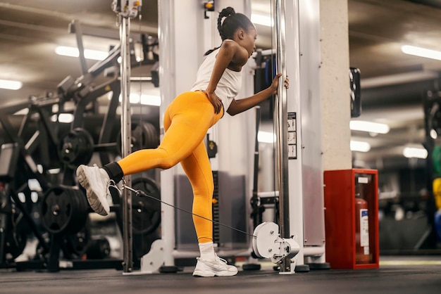 Una magra atleta nera sta praticando i calci su una macchina a doppia puleggia in palestra