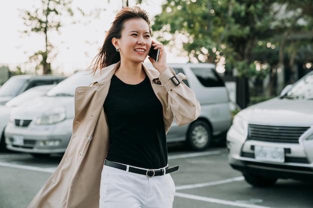 una manager asiatica di successo in smart casual sta camminando vicino a un parcheggio per auto in città mentre fa un piacevole colloquio di lavoro sullo smartphone in una giornata ventosa.