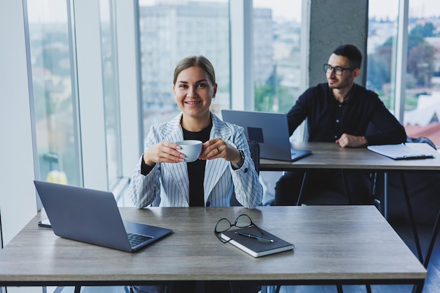 Una manager donna siede alla scrivania utilizzando un computer portatile in un ufficio moderno con un collega sullo sfondo Atmosfera di lavoro in un ufficio con grandi finestre