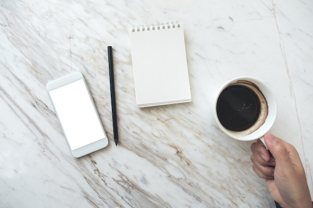 una mano che tiene tazza di caffè con un telefono cellulare con schermo desktop vuoto e notebook vuoto